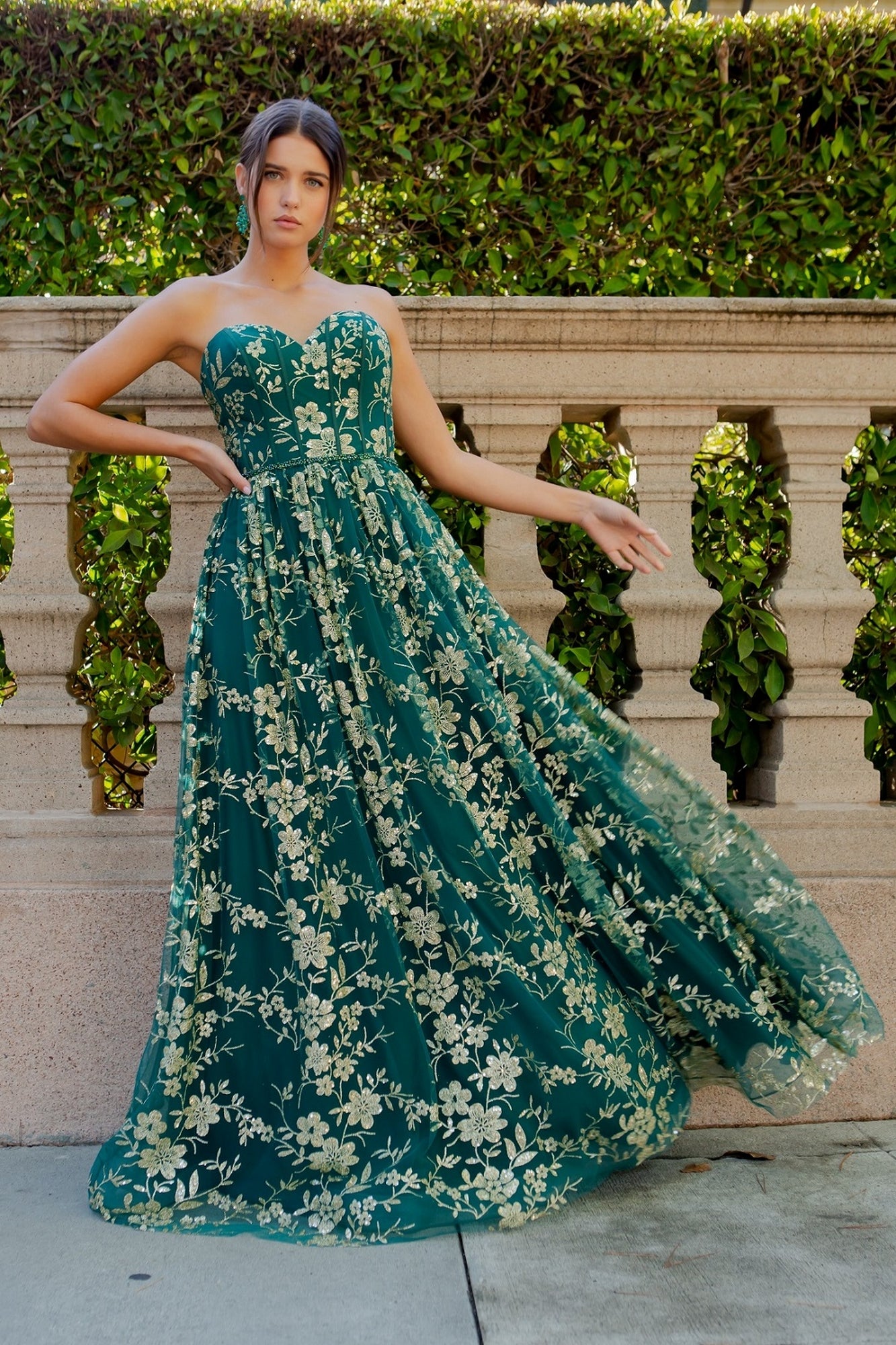 A person wears the Glitter-Floral Long Strapless Prom Dress 8043J, featuring a white glitter floral print, while standing in front of a stone railing and leafy hedge.